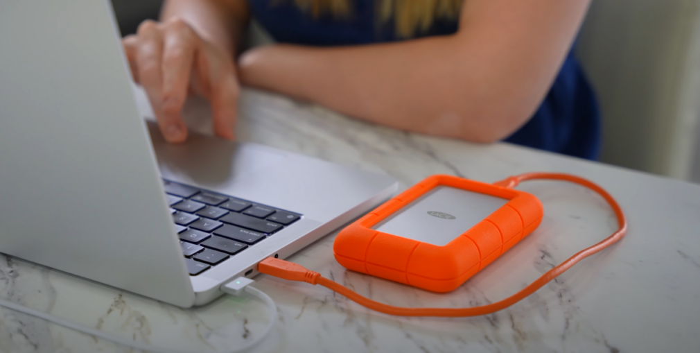 Woman using laptop connected to an orange mini portable hard drive