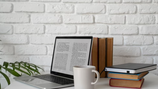 laptop with text, cup, books on the table, and white brick wall behind